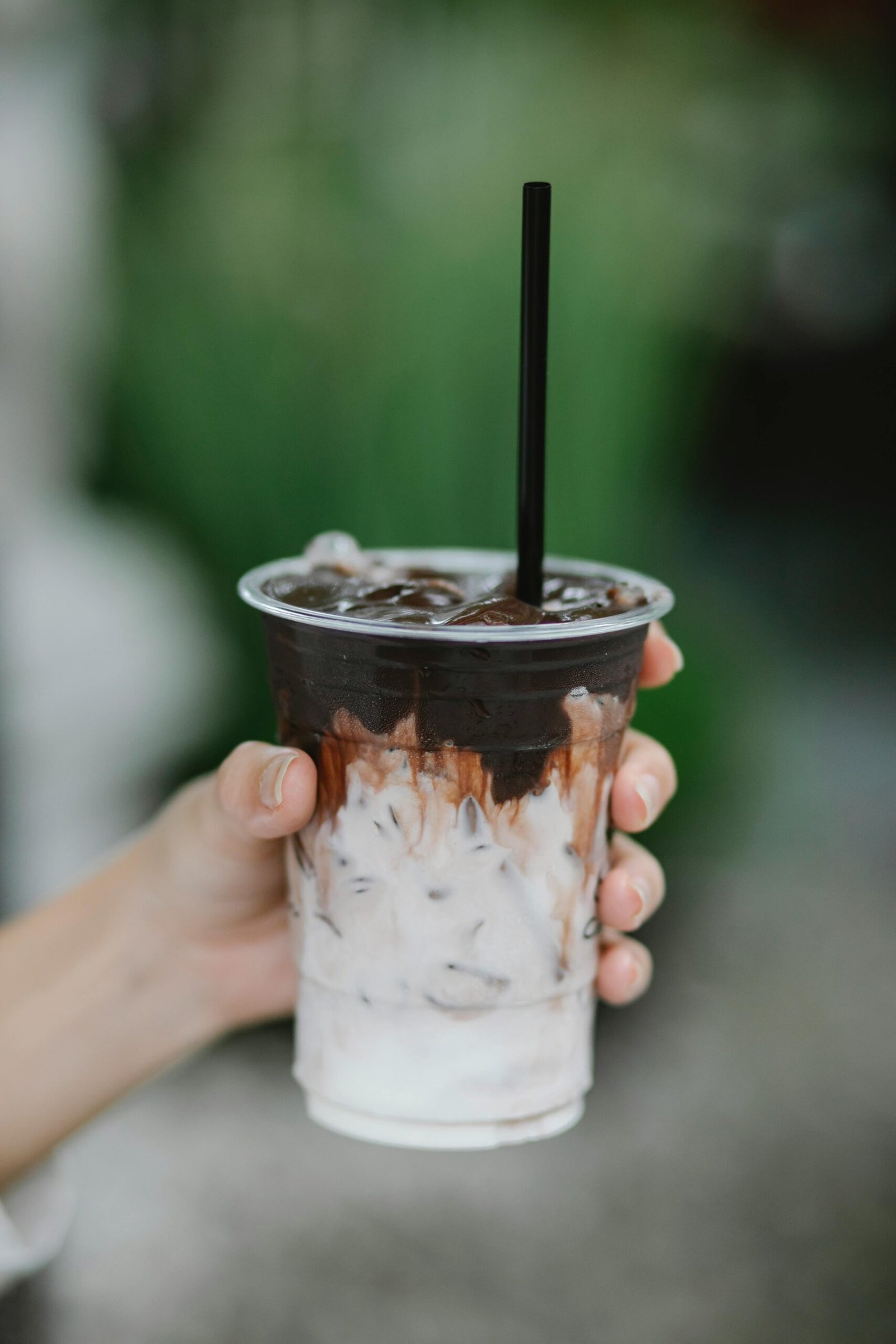 Crop anonymous person with disposable plastic cup of ice coffee with black straw on blurred background